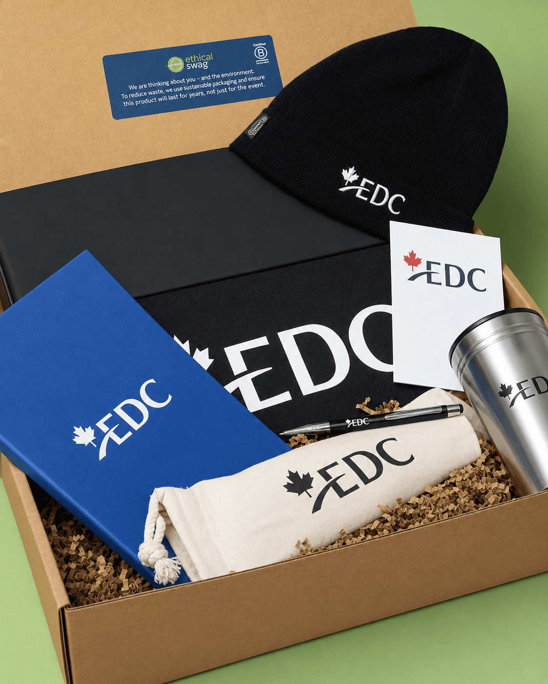 Open EDC employee onboarding kit box with a branded beanie, notebooks, tumbler, pen, and pouch