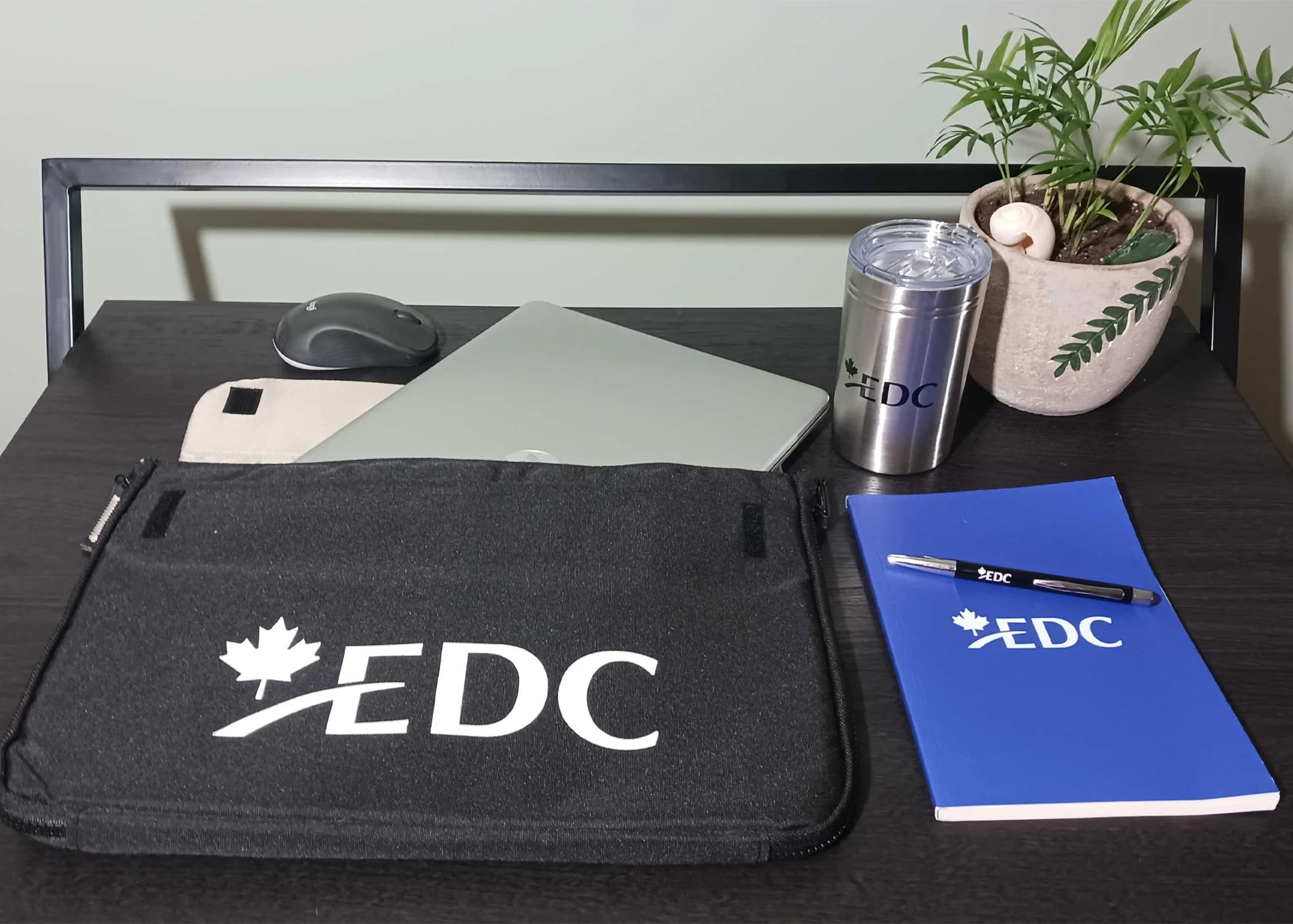 EDC-branded laptop sleeve, tumbler, notebook, and pen on a desk