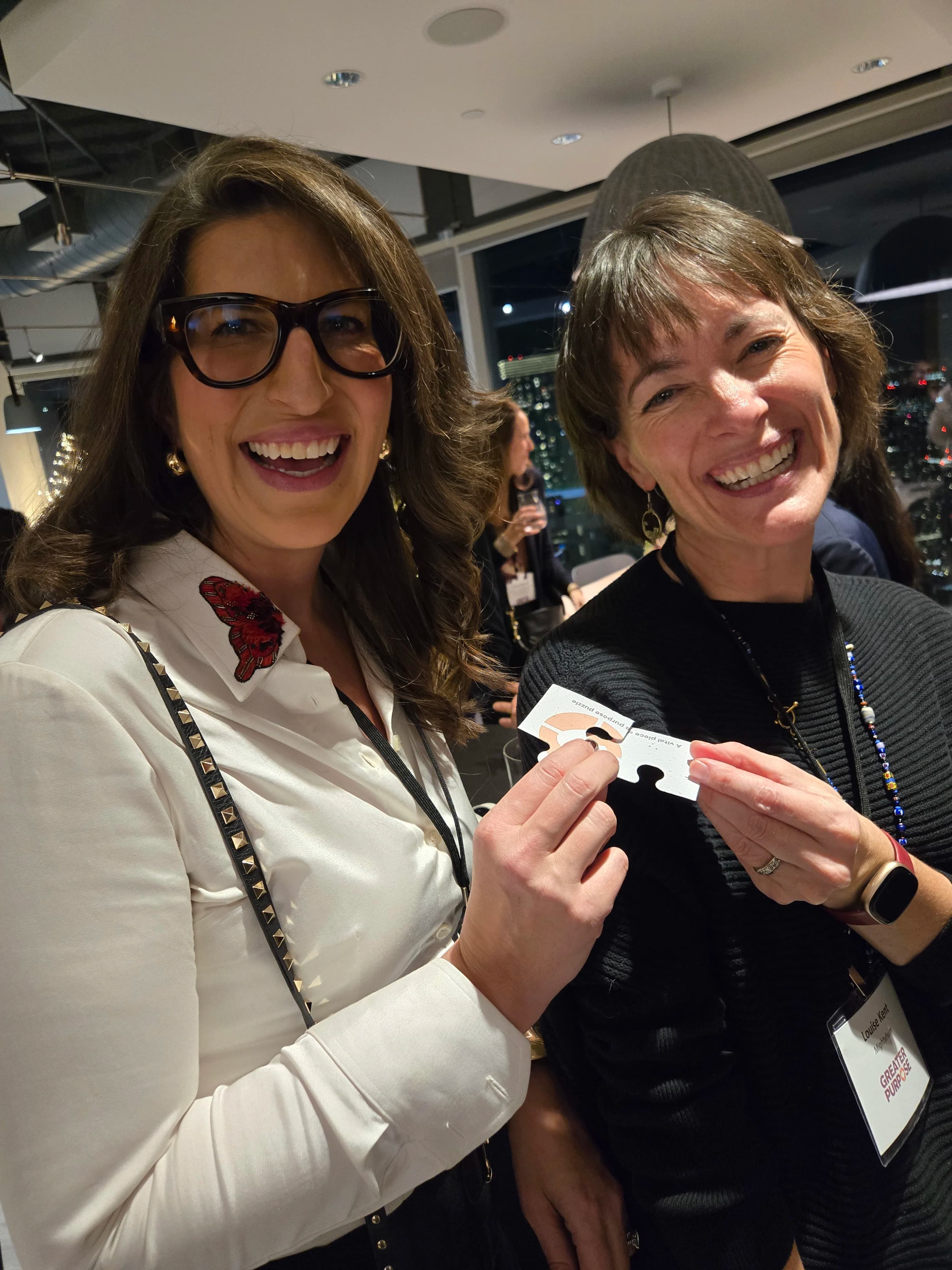 Two attendees at a Greater Purpose networking event laughing and holding branded puzzle-piece cards