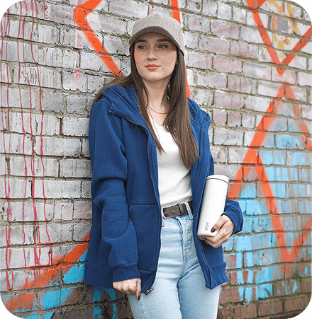 A woman in a blue jacket and white cap standing casually in front of a colorful graffiti wall, representing curated product selection.