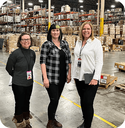 Three smiling women standing in a professional setting, possibly a warehouse or office, suggesting transparent reporting and team collaboration.