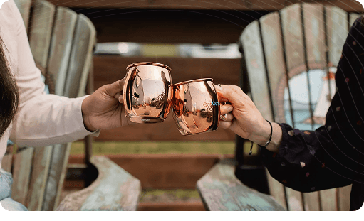 Three individuals' hands holding copper-colored mugs, toasting or sharing a drink in a warm, blurred setting.