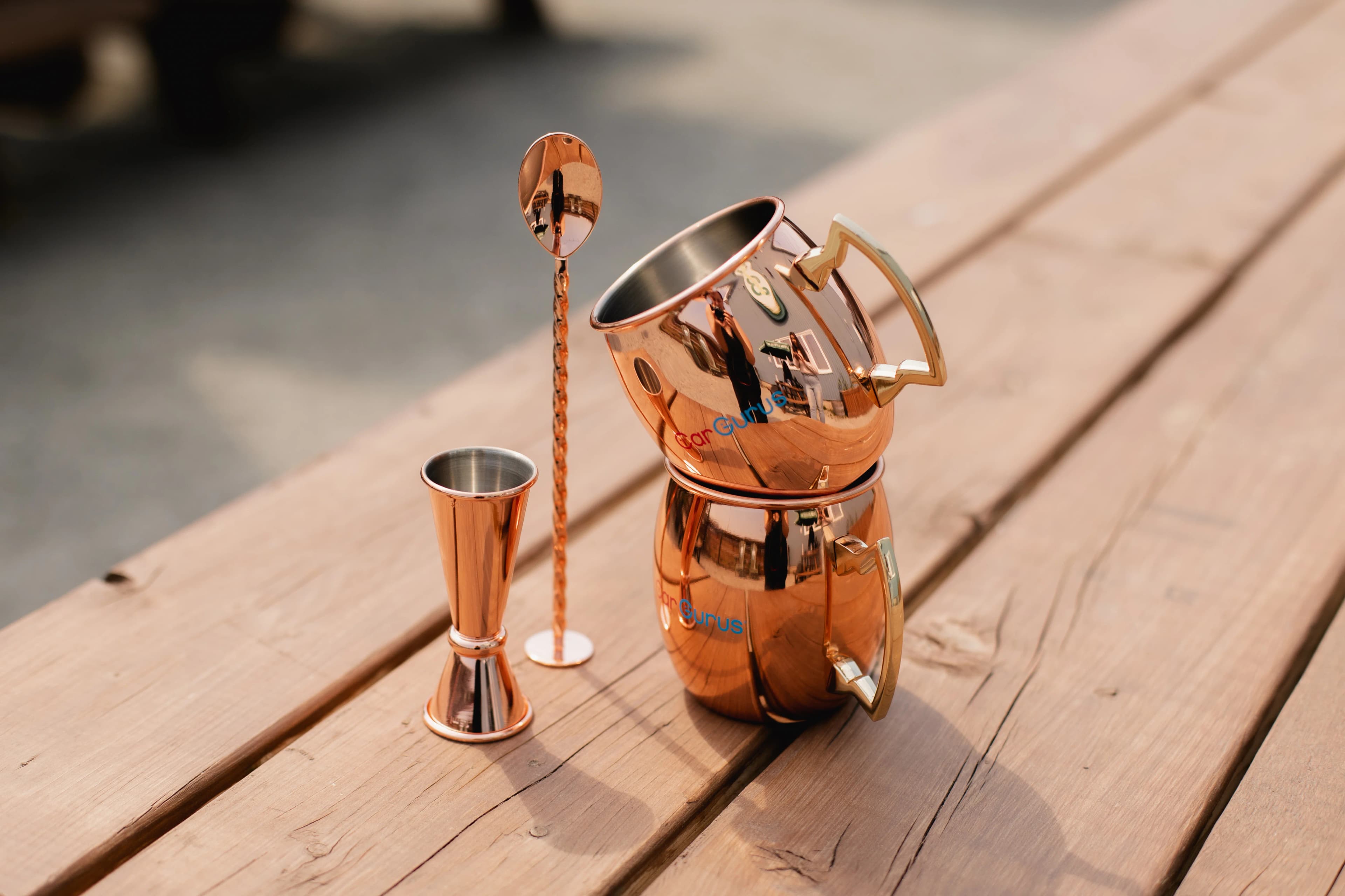 CarGurus-branded copper mug, jigger, and bar spoon set displayed on a wooden bench