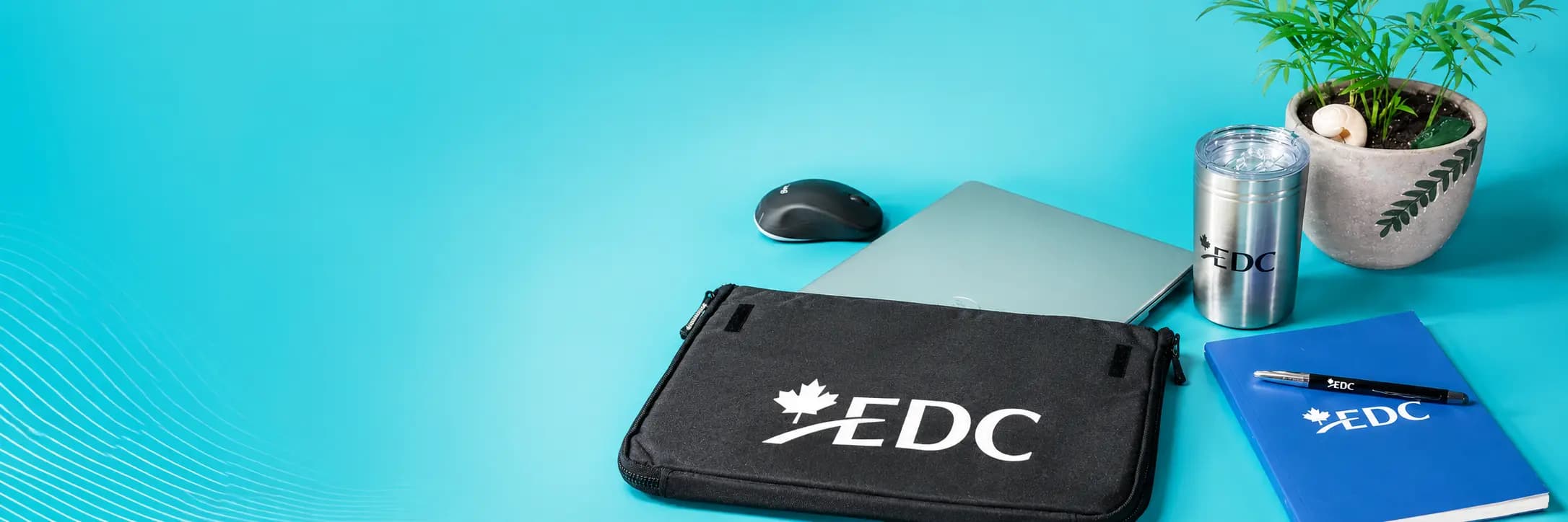 EDC-branded laptop sleeve, tumbler, and notebook arranged next to a laptop on a teal desk backdrop