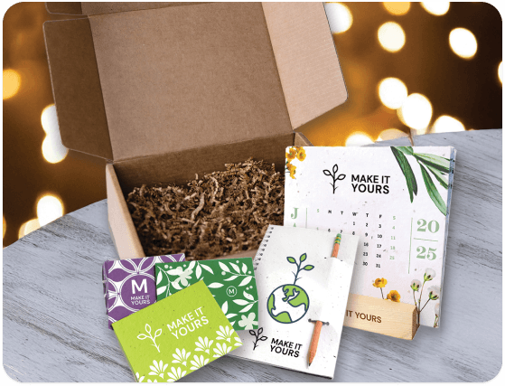 An open brown cardboard box filled with crinkle paper, surrounded by branded, eco-friendly promotional products including a 2025 calendar, a plant-themed notebook with a pencil, and various green and purple cards on a textured light wooden surface with blurred warm lights in the background.